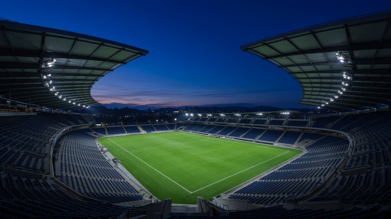 Empty open-air football stadium at blue hour, seen from high in the stands, with a partial roof arching over dark blue seats, a bright green field under the lights and a deep blue Pacific Northwest evening sky