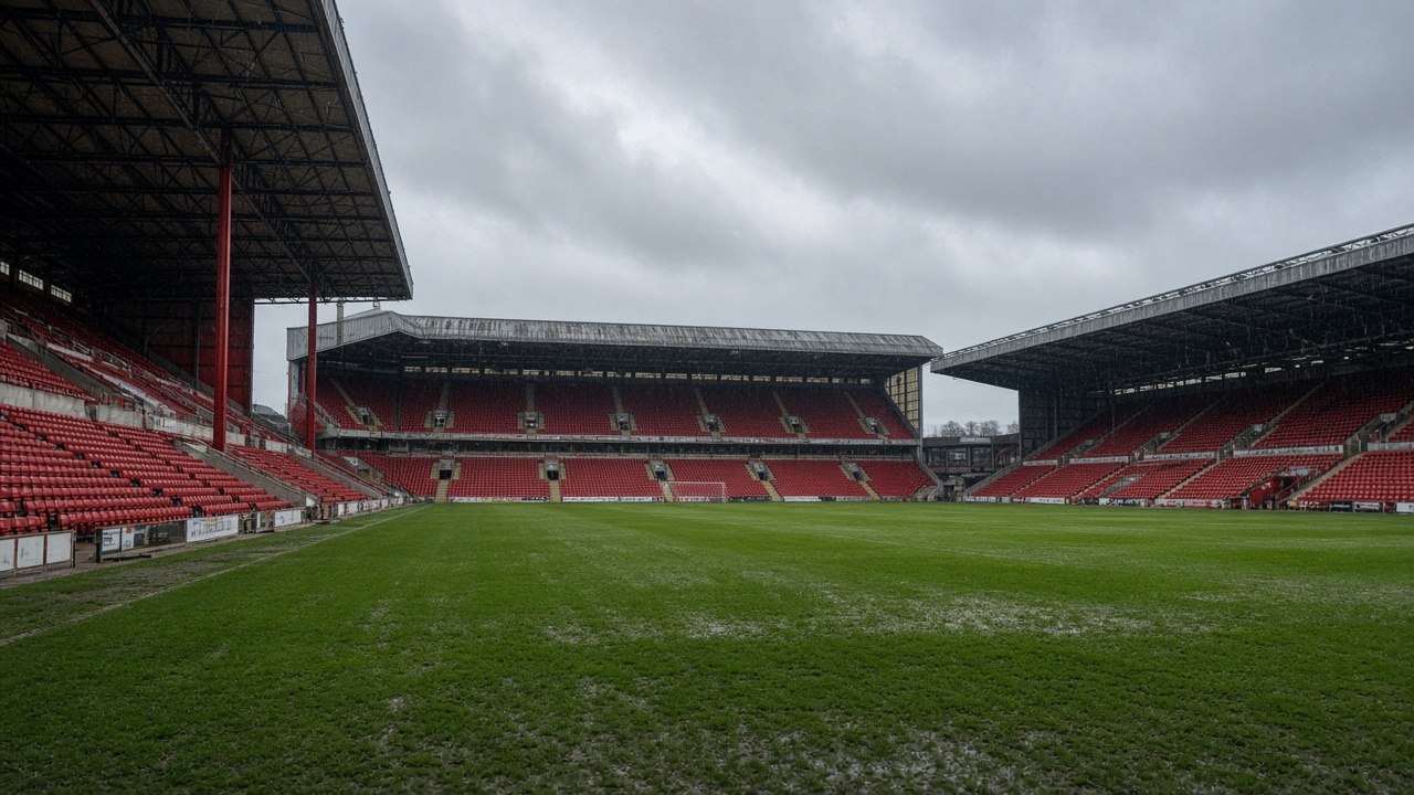 Empty historic football stadium on a grey morning with tall red stands and a green pitch, no people